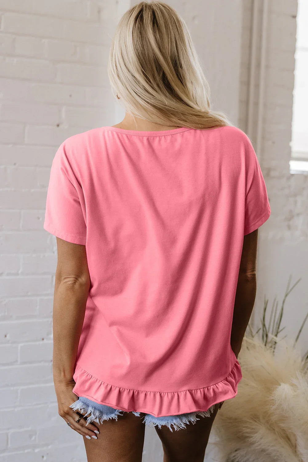 Woman wearing relaxed pink ruffle hem top and denim shorts, standing indoors.