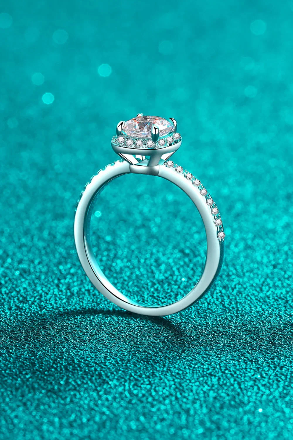 Elegant silver engagement ring with oval diamond center and pavé band on teal glitter background
