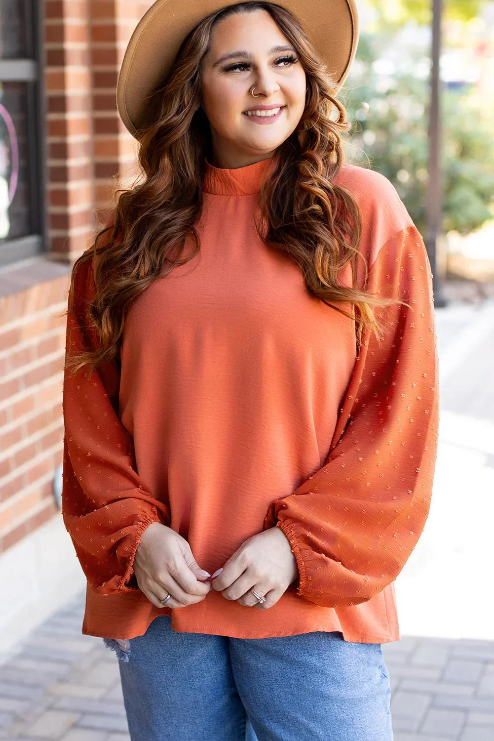 Woman in an orange long sleeve blouse with textured sleeves and blue jeans, outdoors.