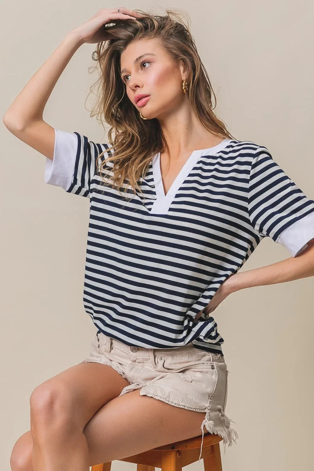 Woman wearing a BiBi Contrast Striped Notched Knit Top with white trim and beige distressed shorts, seated on a stool.