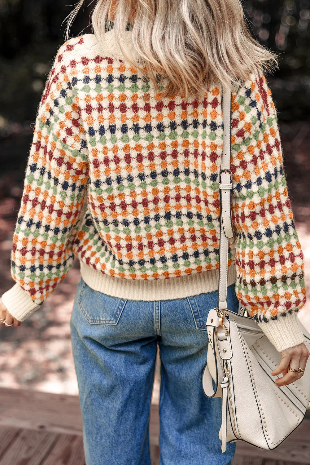 Woman wearing colorful checkered knit sweater, blue jeans, and cream handbag outdoors
