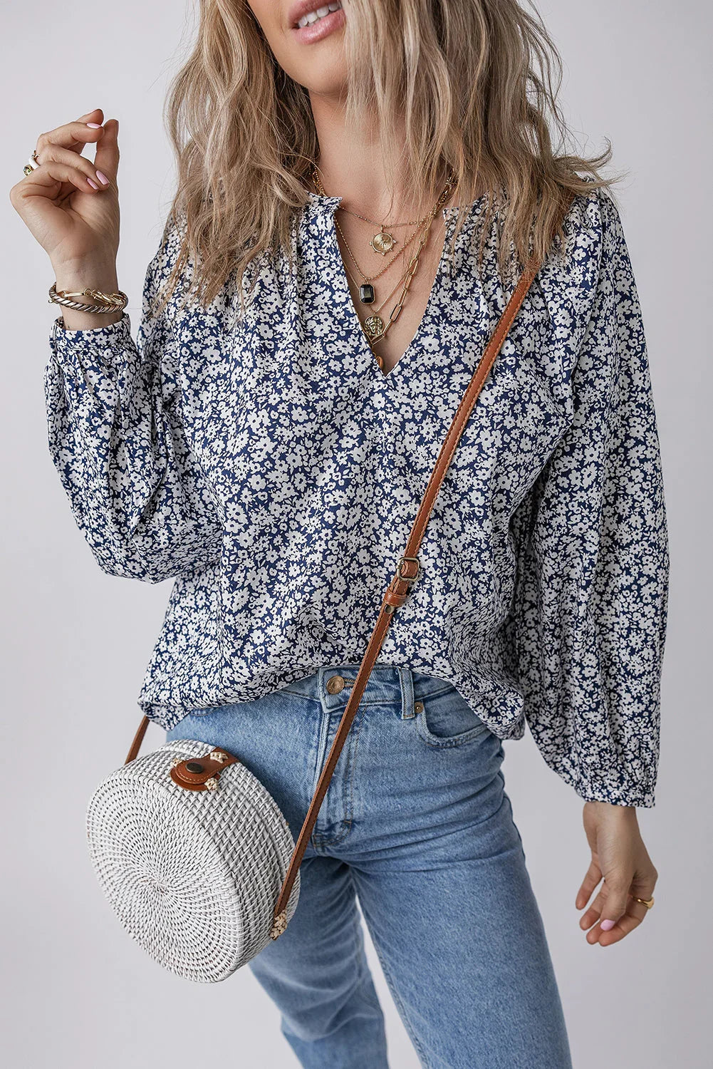 Woman wearing a blue floral blouse, light wash jeans, layered necklaces, and a round woven crossbody bag.