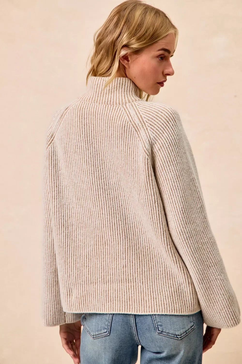 Woman wearing a beige ribbed knit sweater with high neck and light blue jeans, back view
