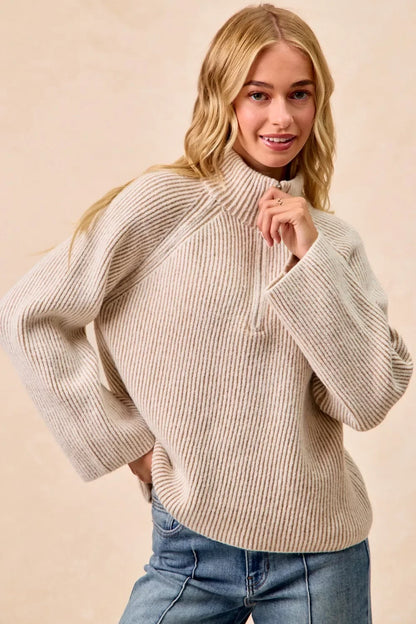 Woman wearing a beige ribbed knit pullover sweater with wide sleeves and a half-zip collar, paired with blue jeans.