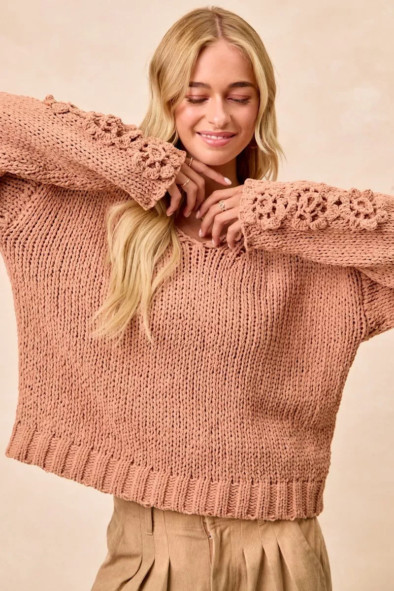 Woman wearing a blush pink chunky knit sweater with crochet floral sleeves and neutral pleated pants.