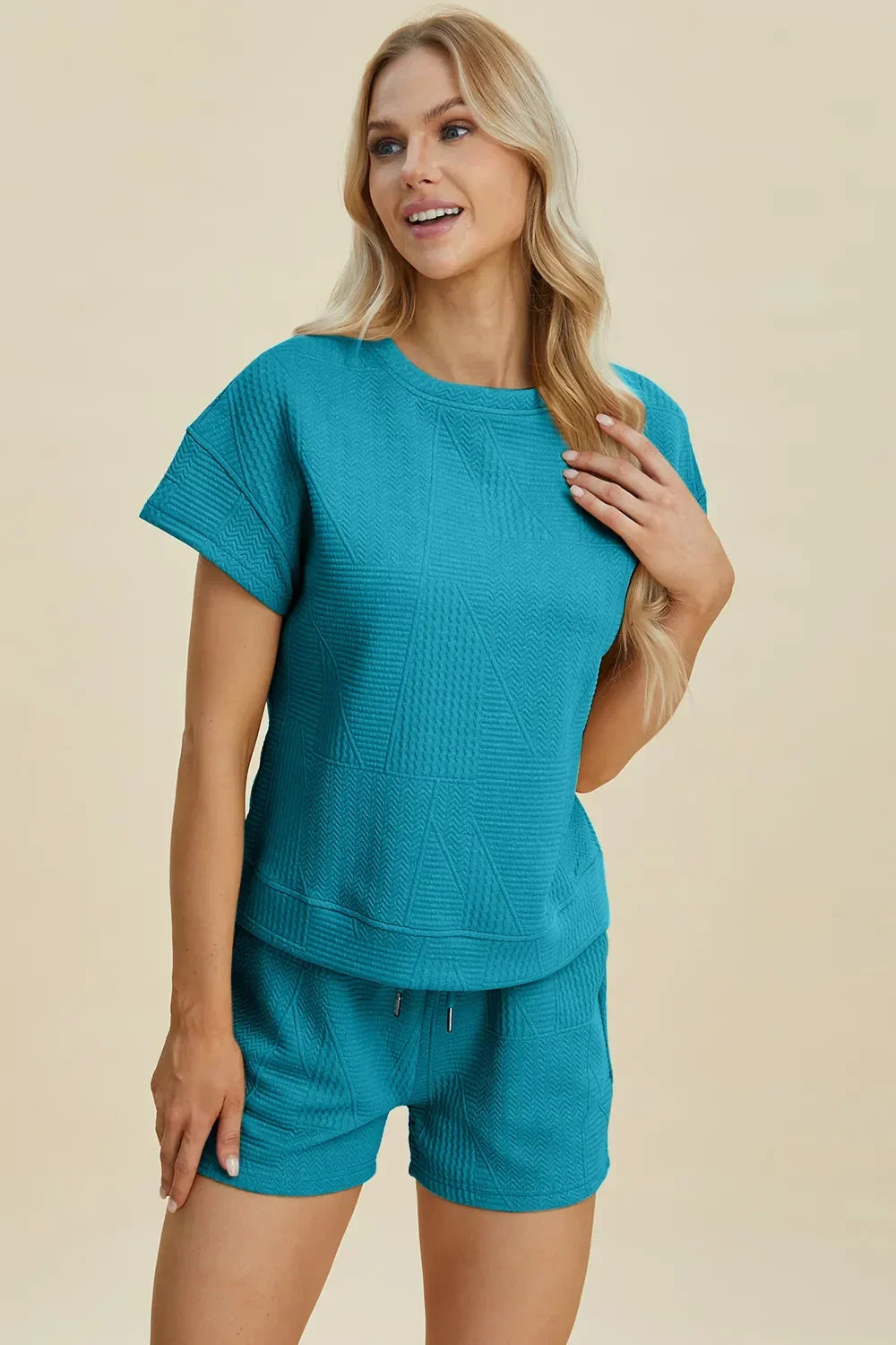 Woman wearing textured teal short-sleeve lounge set with shorts, standing against beige background.