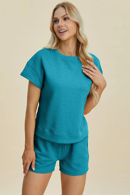 Woman wearing textured teal short-sleeve lounge set with shorts, standing against beige background.