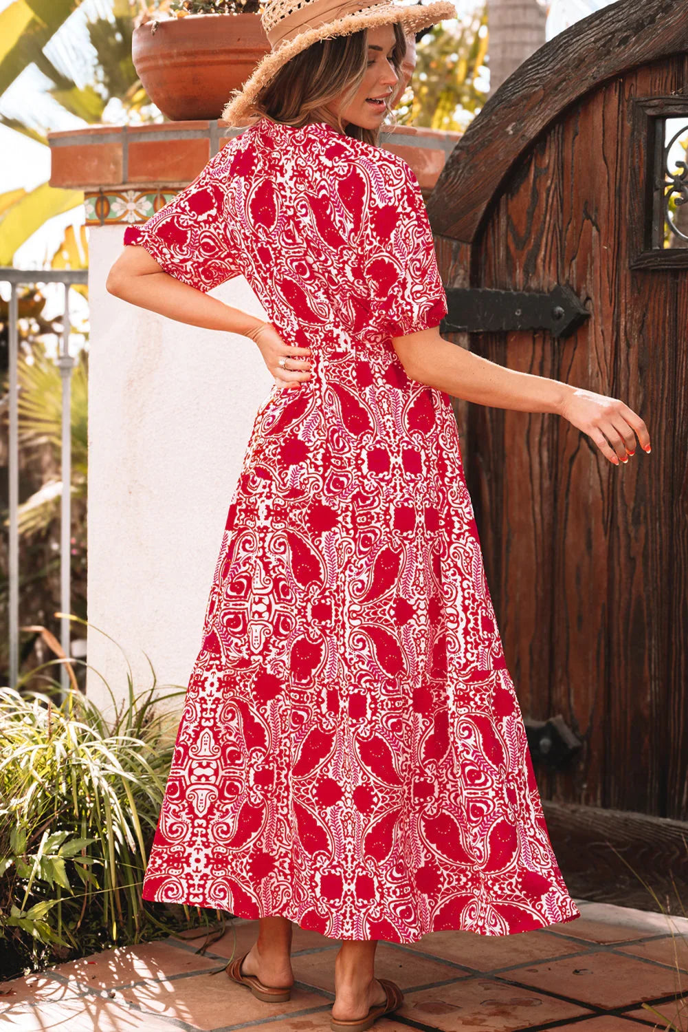 Woman in a red paisley print maxi dress with short sleeves, wearing sandals and a straw hat, standing outdoors by a wooden gate.