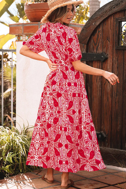 Woman in a red paisley print maxi dress with short sleeves, wearing sandals and a straw hat, standing outdoors by a wooden gate.