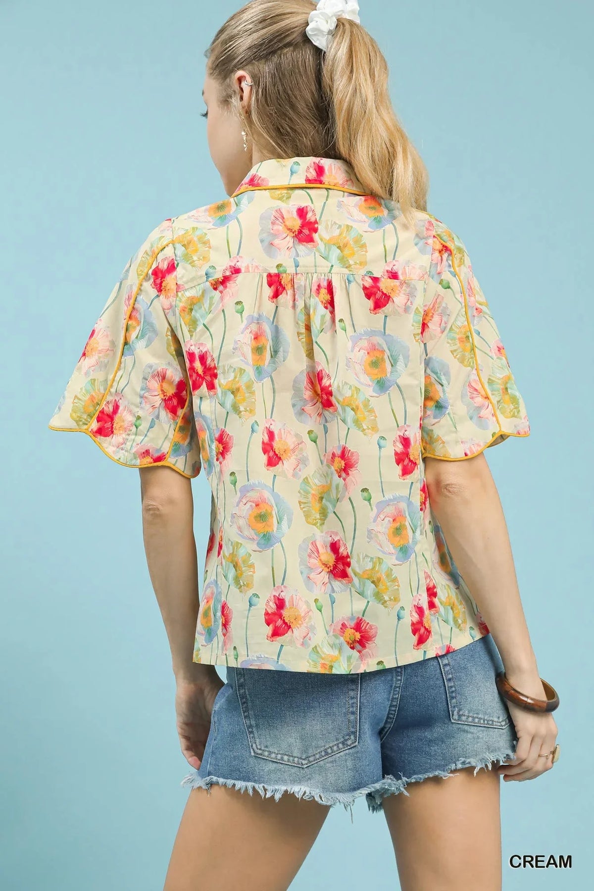 Woman wearing a cream floral short sleeve blouse and denim shorts, shown from the back.