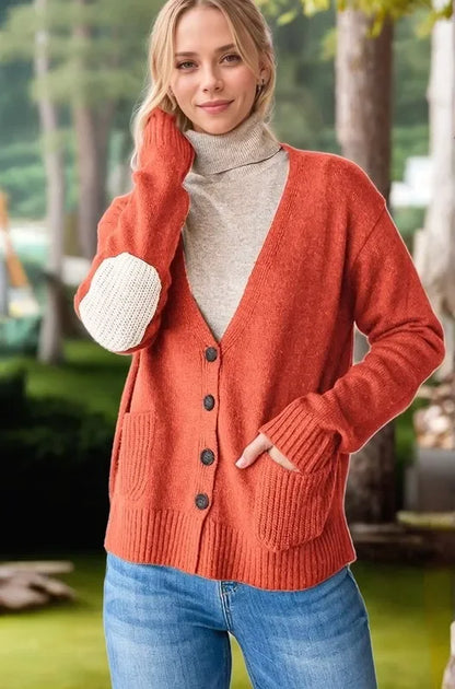 Woman in an orange button-up cardigan with front pockets and white elbow patch, layered over a beige turtleneck, standing outdoors.