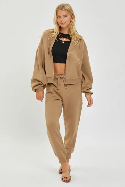 Woman wearing tan sweatsuit with zip-up hoodie, black crop top, and sandals on white background