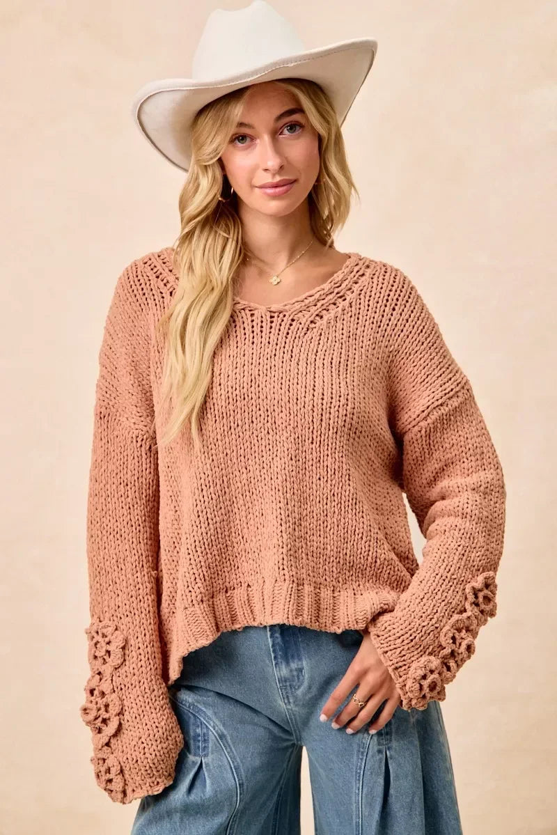 Woman in a white hat wearing a loose knit pink sweater with floral sleeve details and blue jeans