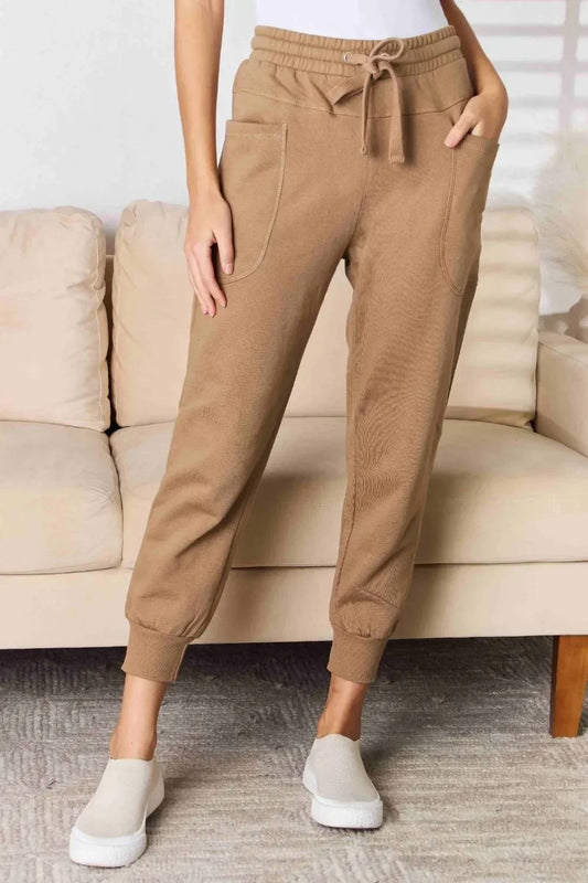 Woman wearing tan jogger pants with drawstring waist, front pockets, and slip-on shoes indoors