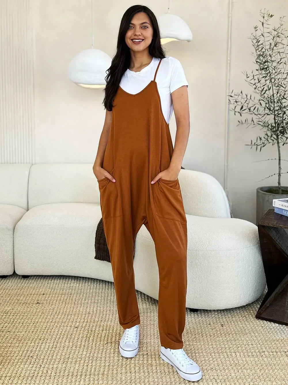 Woman wearing rust brown relaxed jumpsuit over white t-shirt and sneakers in modern living room