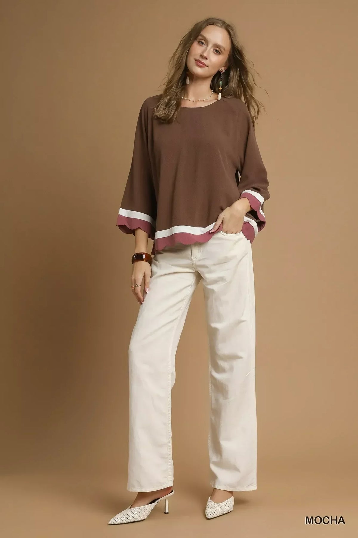 Woman in a relaxed mocha brown top with flared sleeves, white pants, and white heels on neutral background.