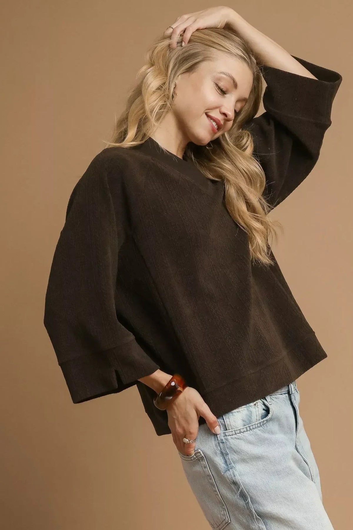 Woman wearing loose dark brown top with wide sleeves and light wash jeans, neutral background