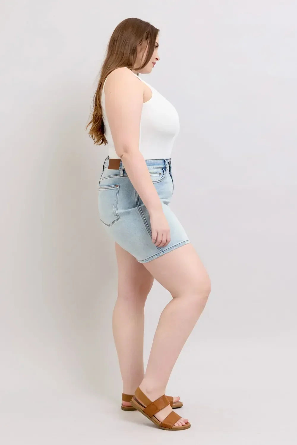 Woman in light wash denim shorts, white sleeveless top, and tan sandals, side view