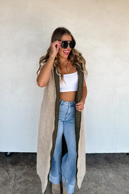 Woman in long sherpa vest, white crop top, wide-leg jeans, and sunglasses leaning against wall