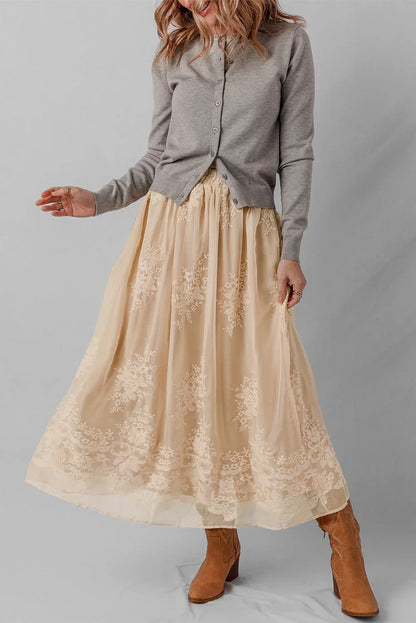 Woman wearing a grey cardigan, beige embroidered maxi skirt, and tan suede boots.