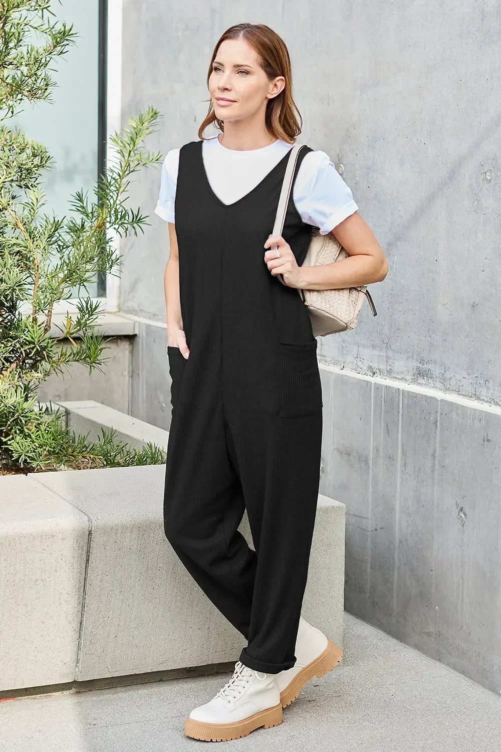 Woman in a black relaxed jumpsuit and white t-shirt, standing outdoors with a tan bag and boots