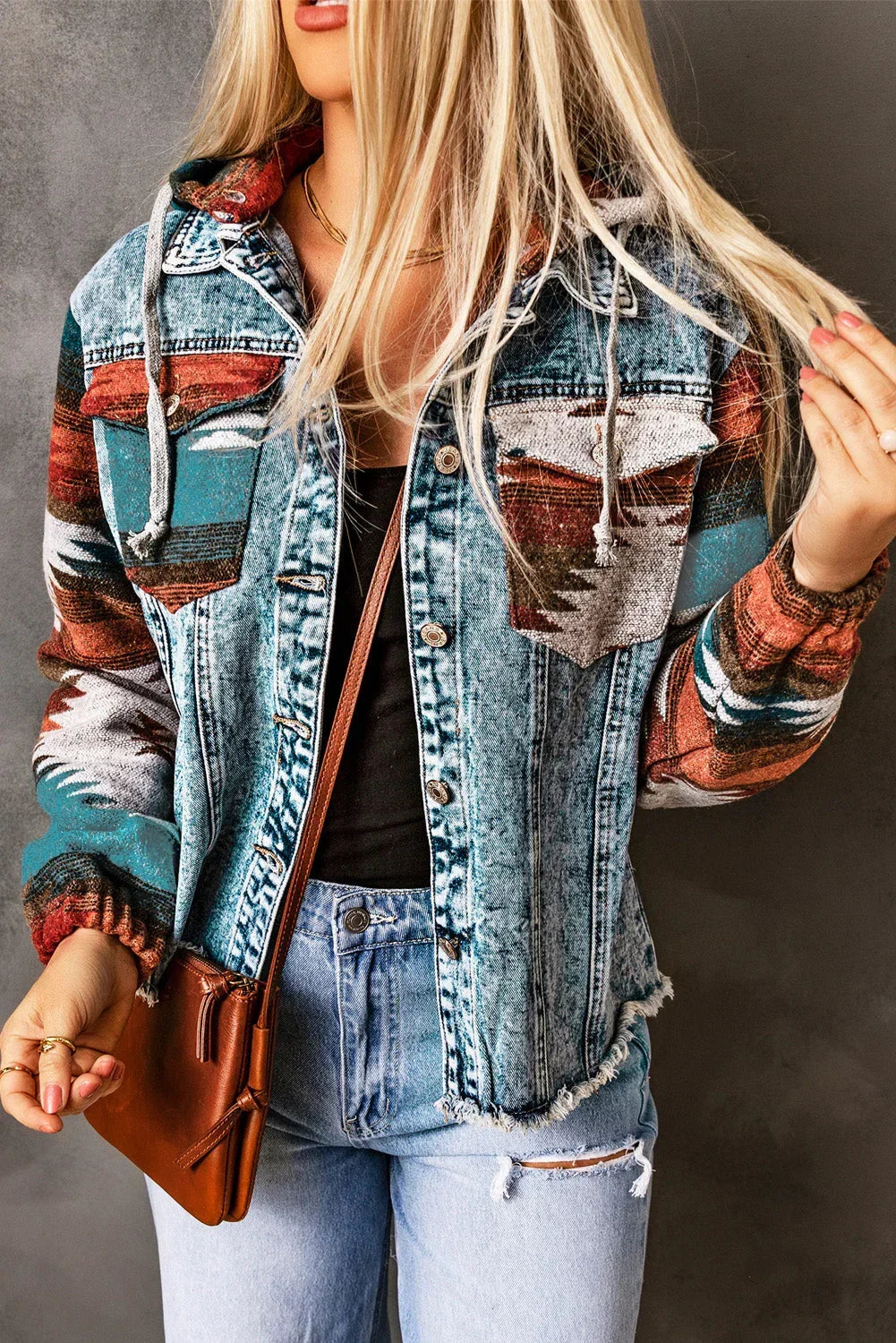 Woman wearing a distressed denim jacket with colorful patterned sleeves, light wash jeans, and a brown crossbody bag.