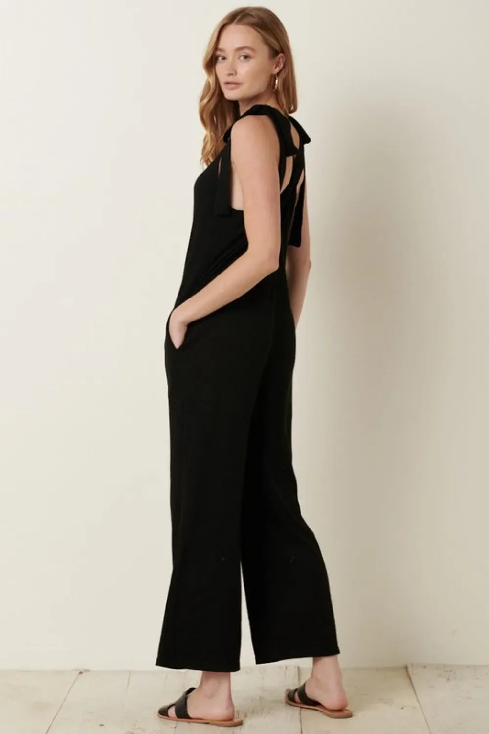 Woman wearing a sleeveless black jumpsuit with tie shoulder straps and black sandals.