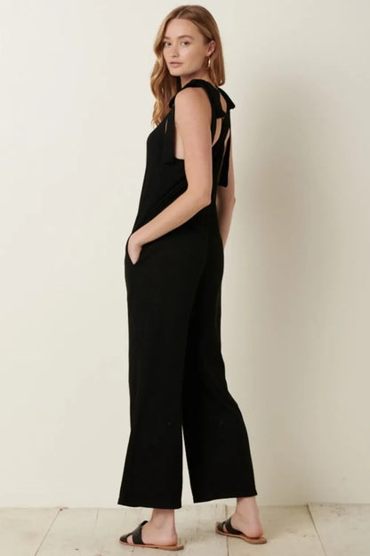 Woman wearing a sleeveless black jumpsuit with tie shoulder straps and black sandals.