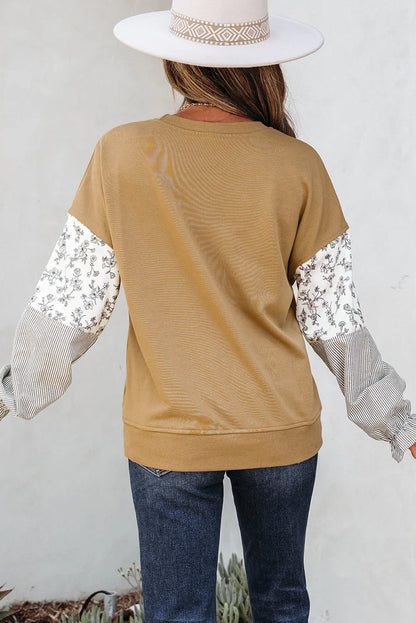 Woman wearing a camel sweater with floral and striped patchwork sleeves, blue jeans, and a white hat