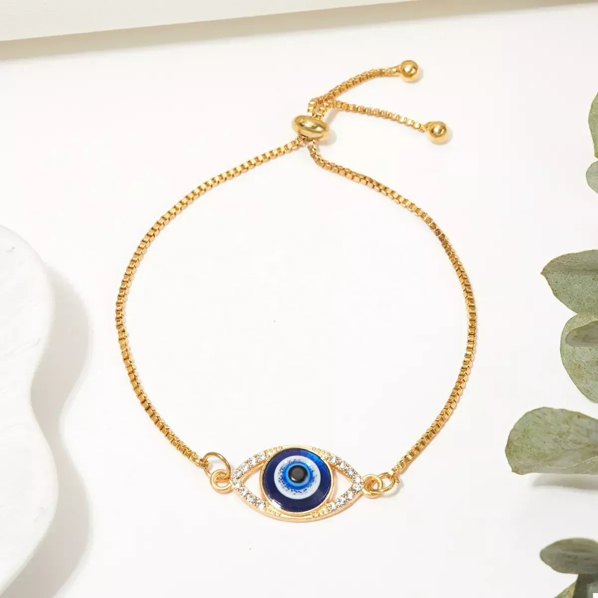Gold adjustable bracelet with blue evil eye charm and clear crystal accents on white background