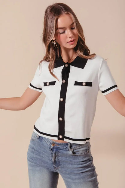 Woman wearing a short sleeve white knit top with black trim, pearl buttons, and blue jeans