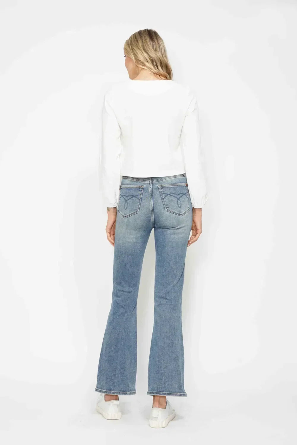 Woman wearing light wash flare jeans, white long sleeve top, and white sneakers, back view