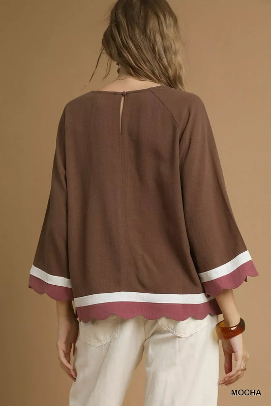 Back view of a woman wearing a loose mocha brown top with scalloped hem and white trim, paired with light pants.