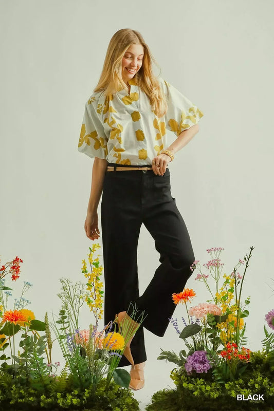 Woman in wide-leg black pants and a white floral blouse standing among colorful flowers.