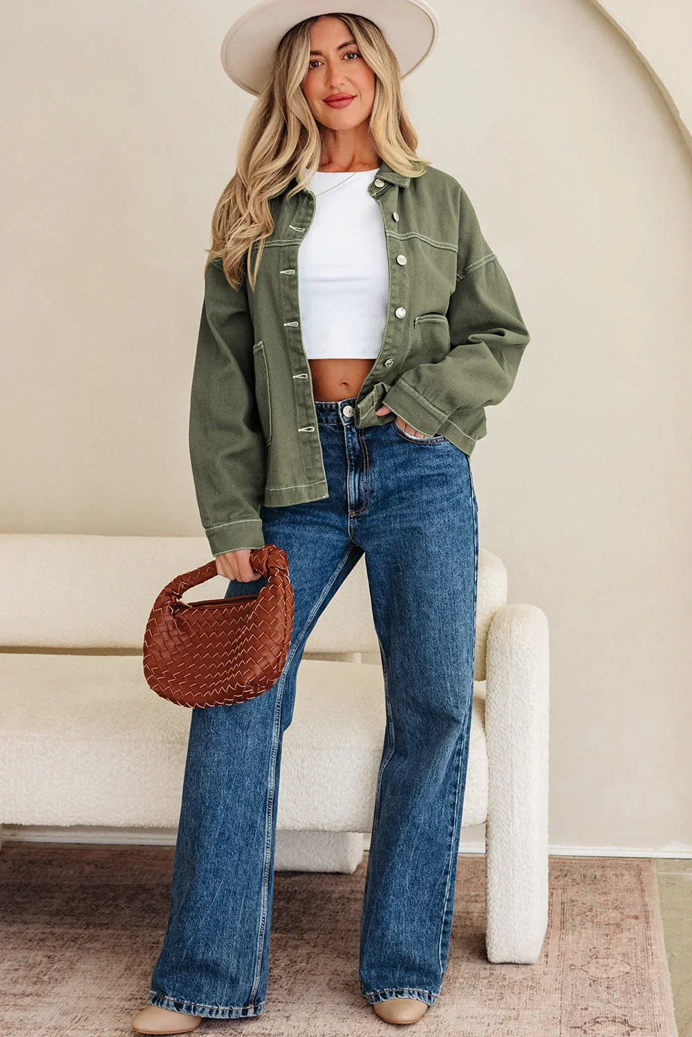 Woman in olive green jacket, white crop top, wide-leg jeans, and hat holding brown woven bag