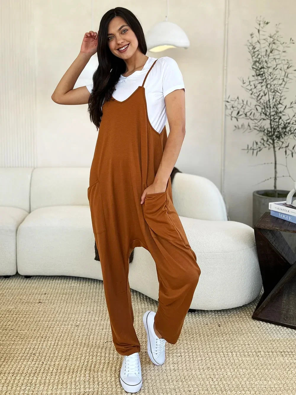 Woman wearing relaxed brown jumpsuit over white t-shirt with white sneakers in modern living room.