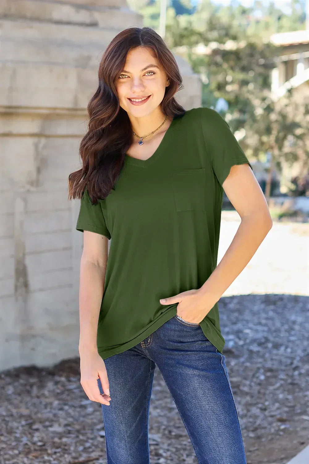 Woman wearing an olive green short sleeve V-neck top with front pocket and blue jeans outdoors.