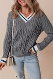 Woman wearing a gray cable knit sweater with white and blue trim, holding a beige handbag against a beige background.