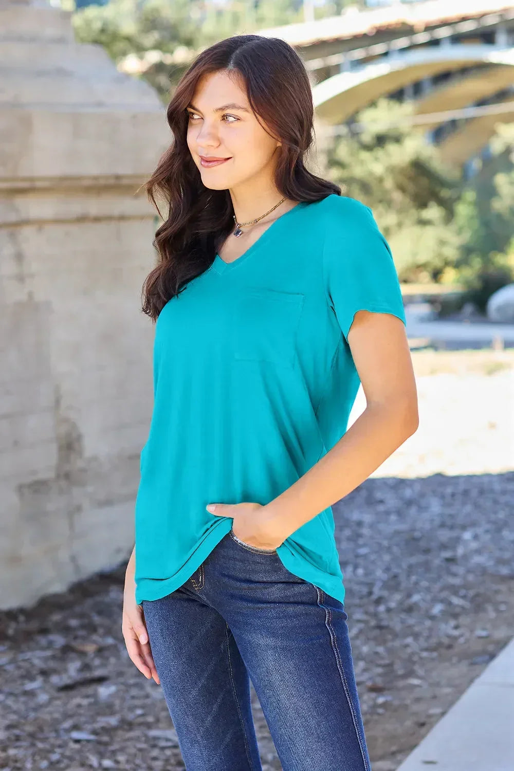 Woman wearing a turquoise V-neck short sleeve top with front pocket and blue jeans outdoors.