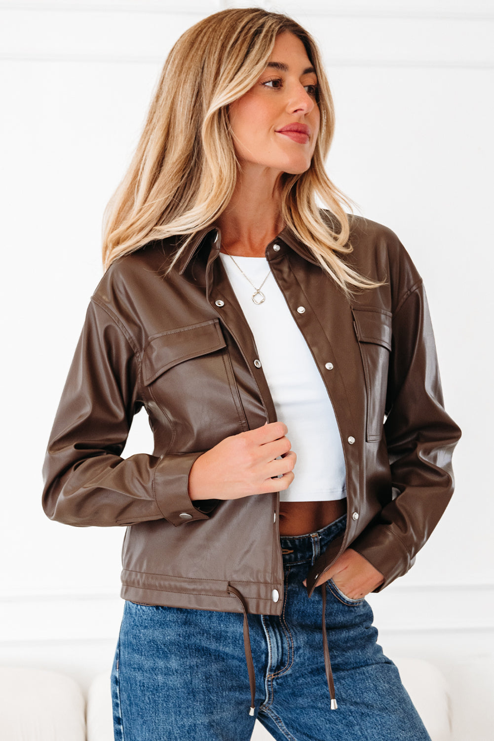 Woman wearing a brown faux leather jacket, white crop top, and blue jeans, standing indoors.