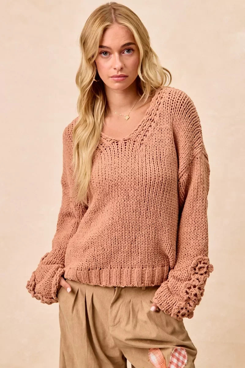 Woman wearing a relaxed, taupe knit sweater with floral sleeve details and tan pleated pants.