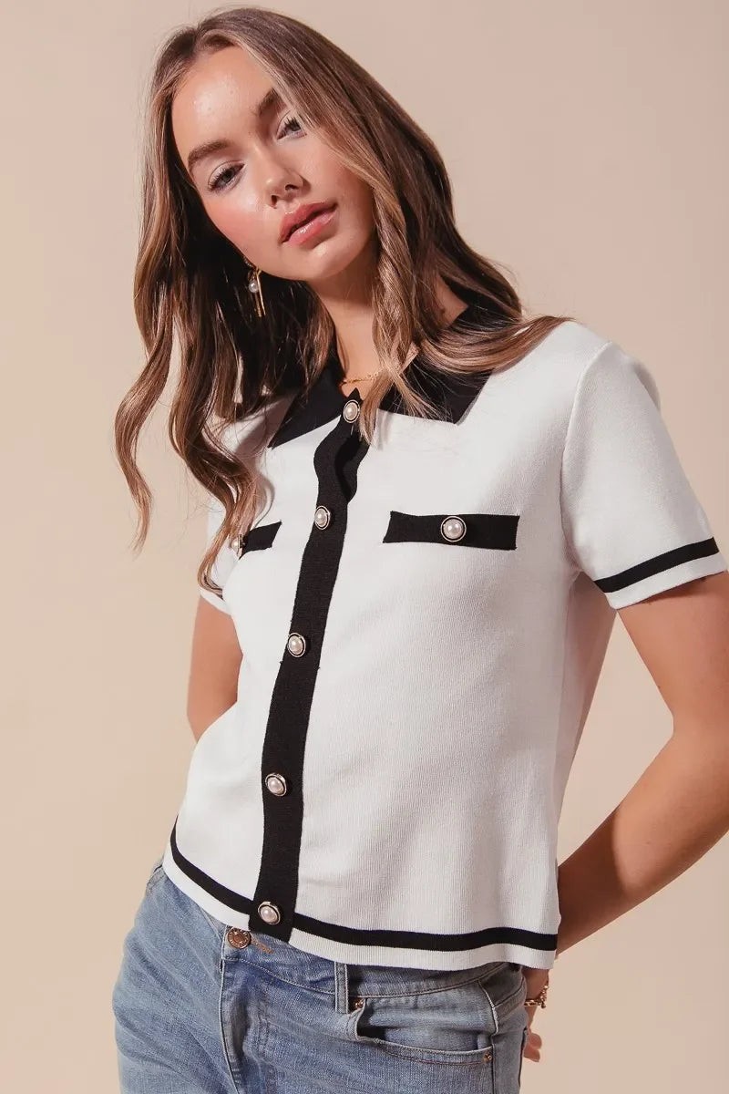 Woman wearing a short-sleeve white knit top with black trim and pearl buttons, paired with jeans.