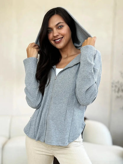 Woman wearing a light grey ribbed zip-up hoodie with hood up, in a modern indoor setting.