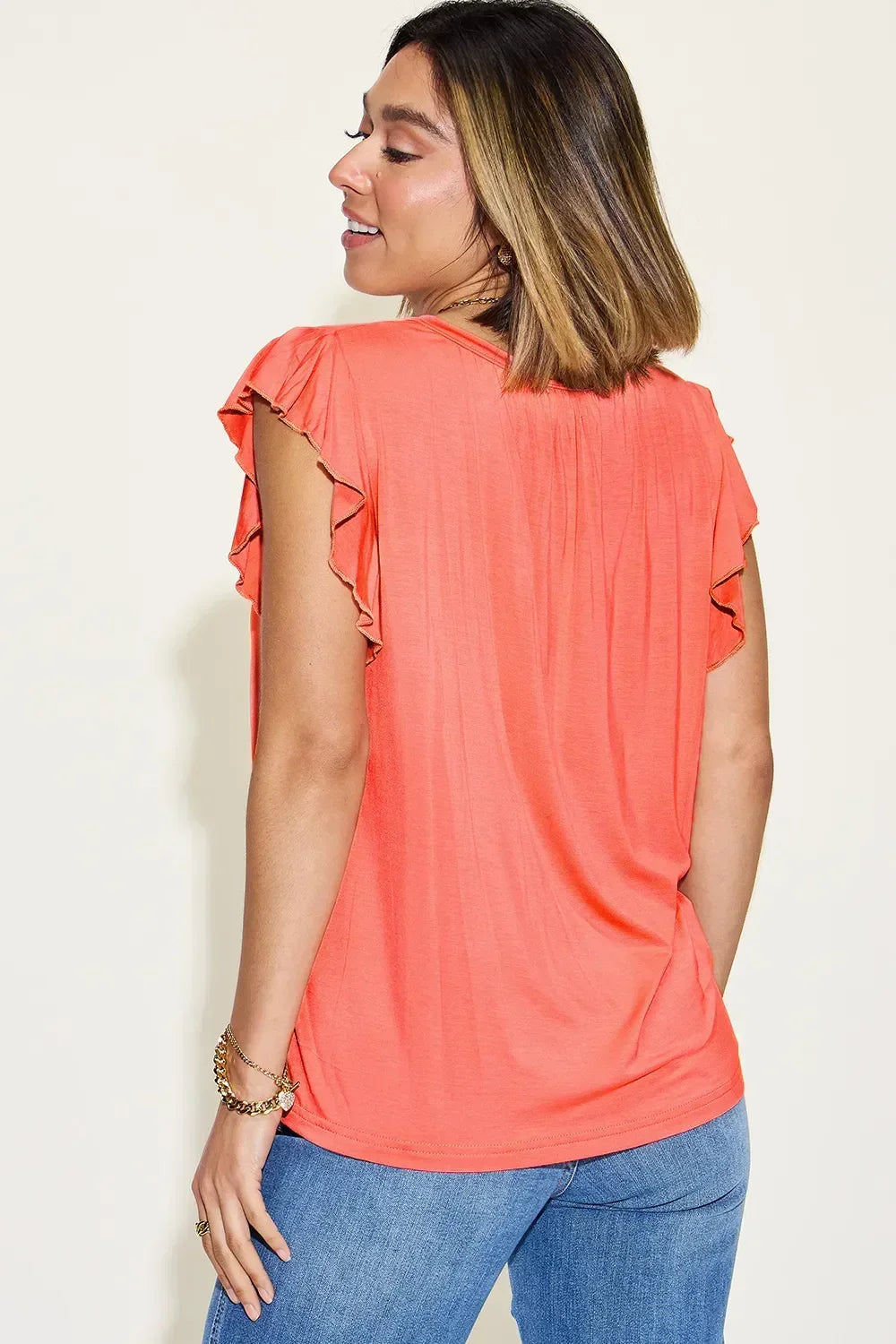 Woman wearing coral flutter sleeve top and blue jeans, shown from the back