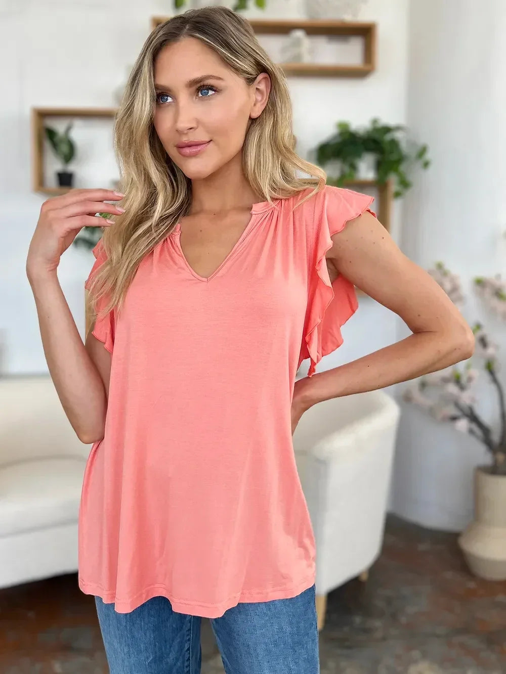 Woman wearing a coral ruffle sleeve top and blue jeans in a modern living room setting.