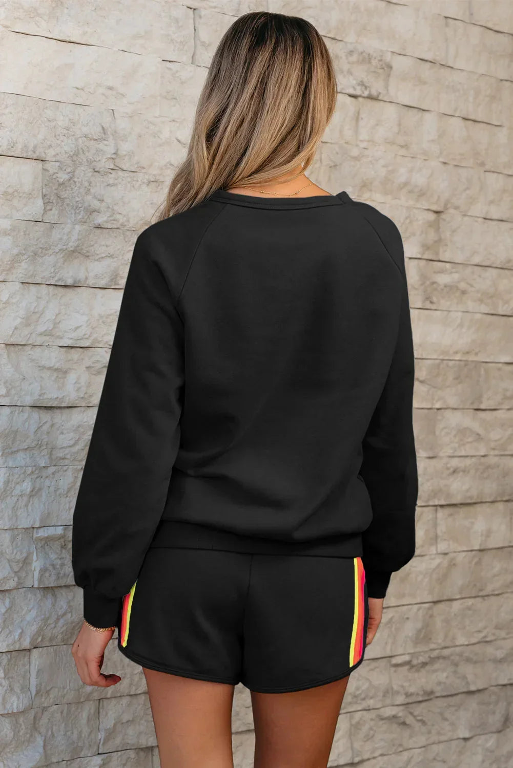 Woman wearing black long sleeve top and black shorts with side stripe, standing by stone wall