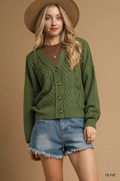 Woman in olive green cable knit cardigan, denim shorts, and tan hat against beige background
