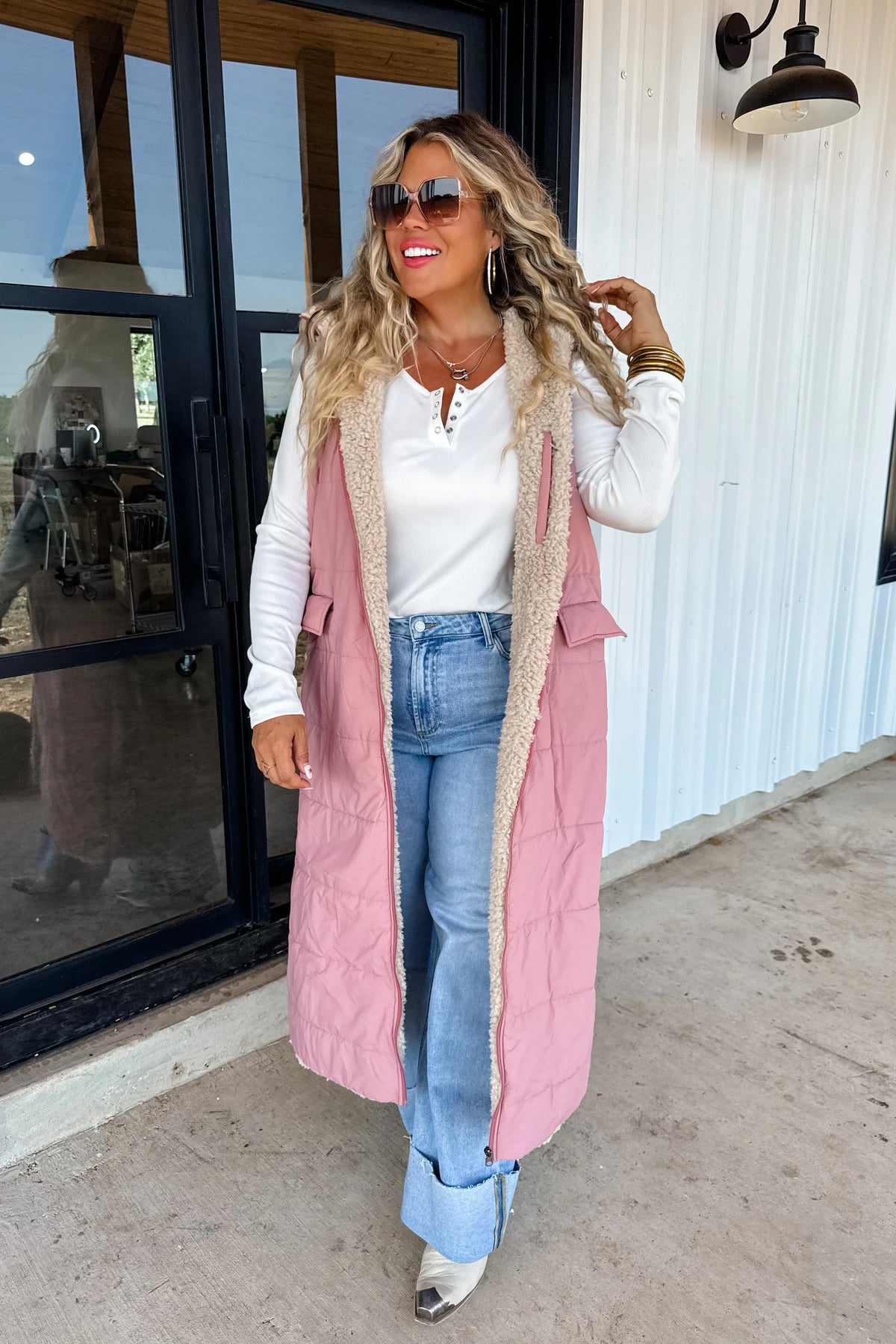 Woman wearing long pink sherpa-lined vest, white henley top, light blue wide-leg jeans, and sunglasses