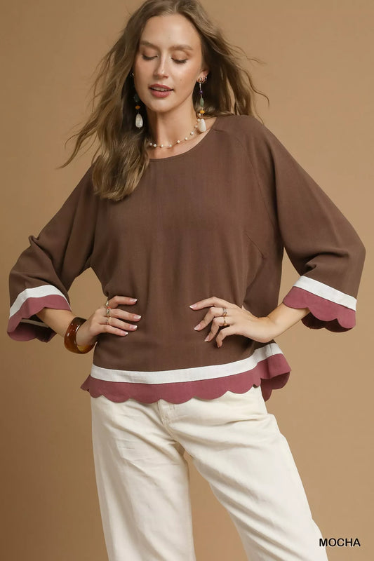 Woman wearing a brown scalloped crewneck blouse with white pants against a beige background.