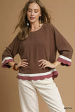 Woman wearing a brown scalloped crewneck blouse with white pants against a beige background.